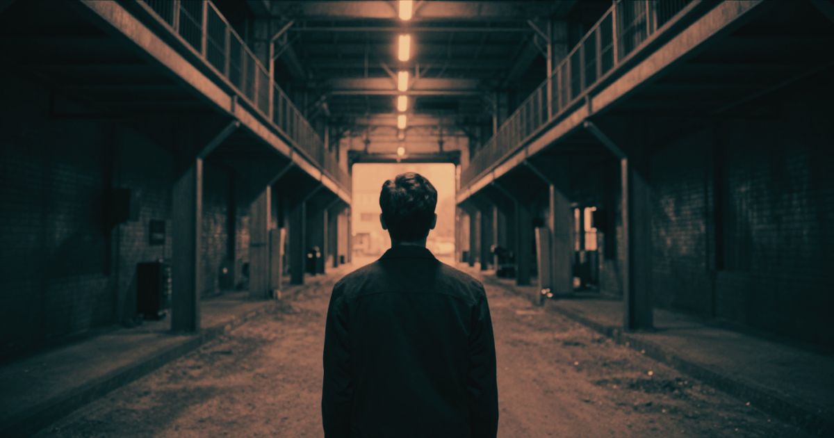A lone figure seen from behind at the mouth of two near-identical corridors, one leading into depth and one a dead end