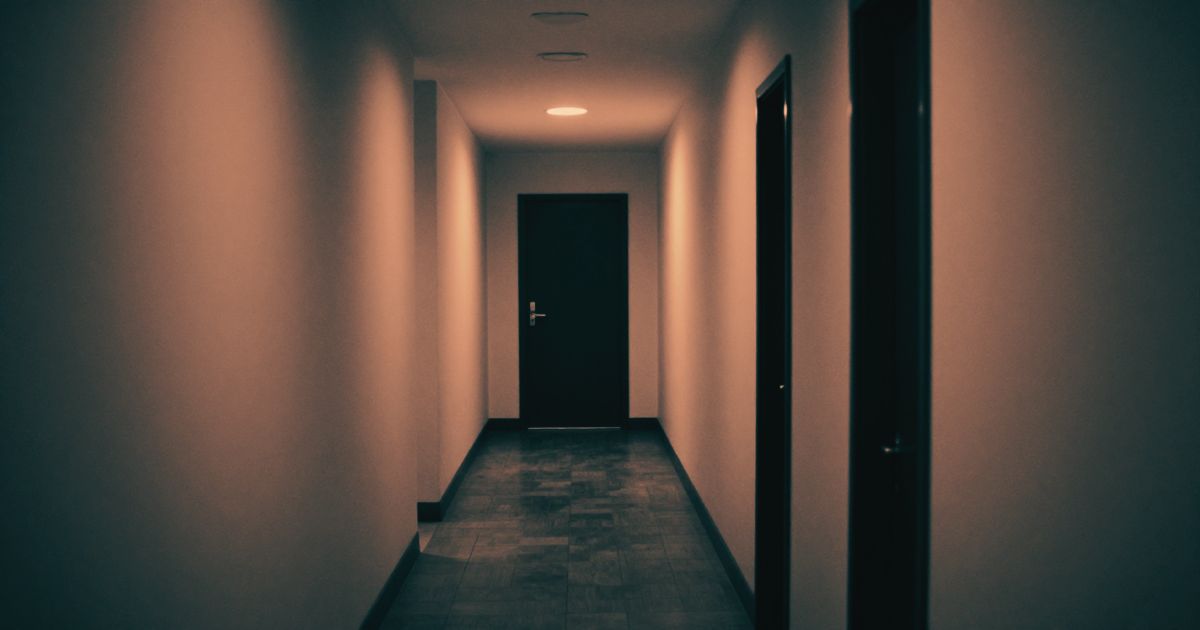 A long empty corridor with a single door at the far end, shot from floor level, high contrast