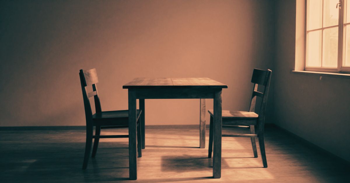 Two chairs facing each other across a bare wooden table, empty room, high window light from one side