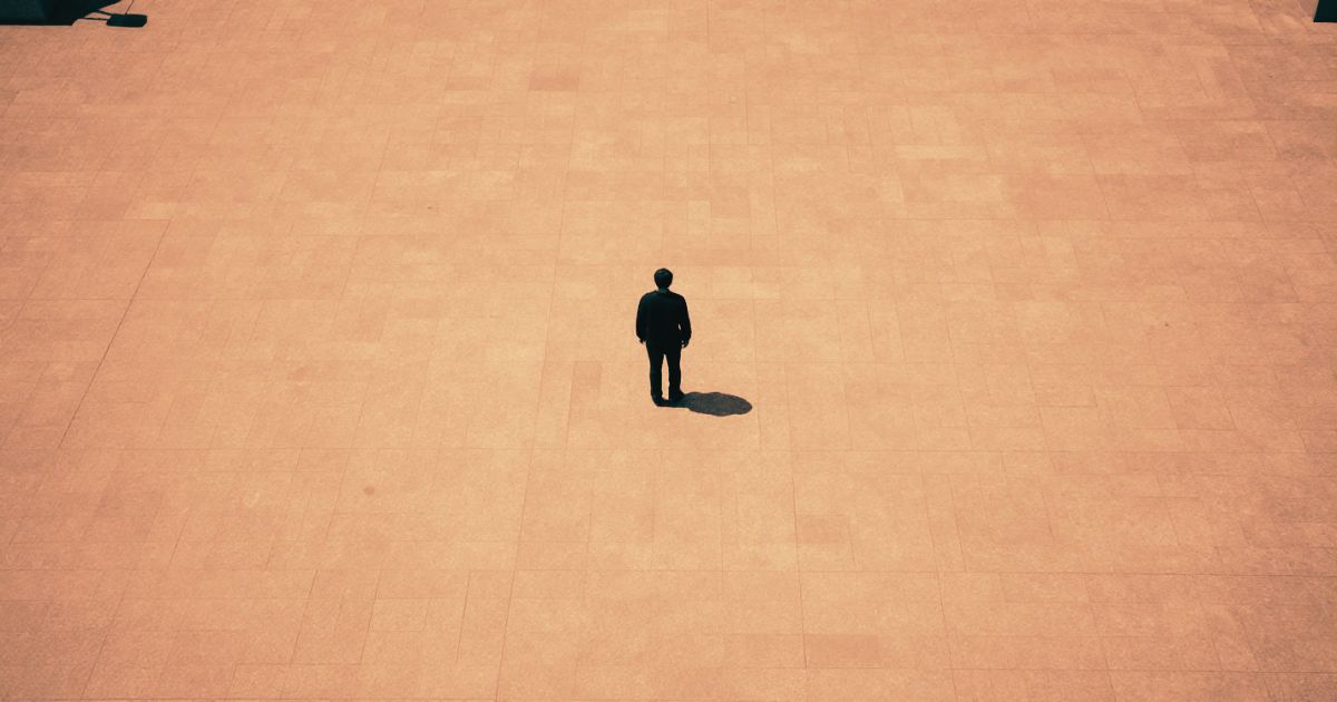 A lone figure standing at the centre of a vast empty public square, shot from above, dwarfed by the space around them