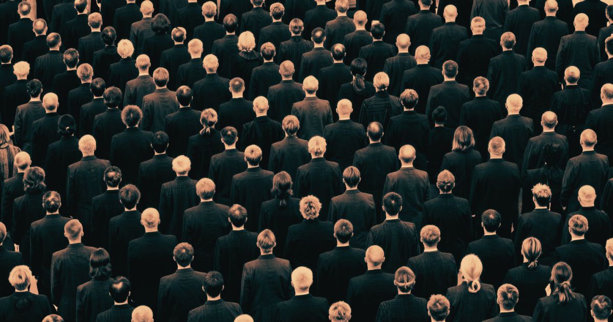 An enormous crowd of identical suited figures viewed from above, one figure standing slightly apart from the mass