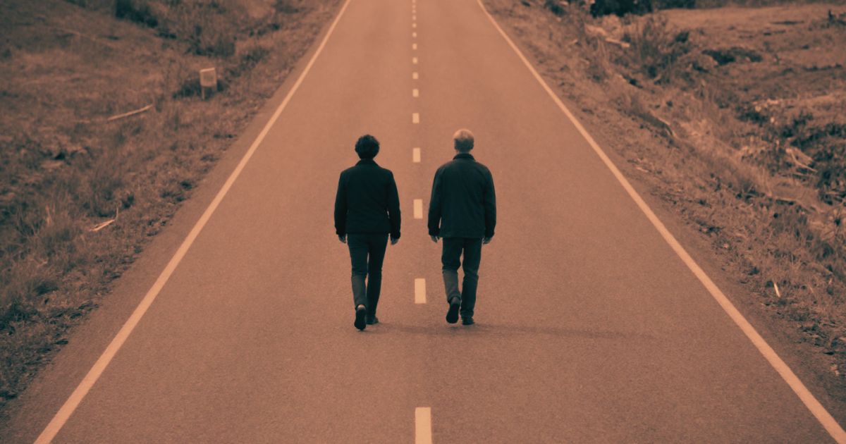 Two figures walking toward each other along a long empty road, shot from above, vast empty space around them
