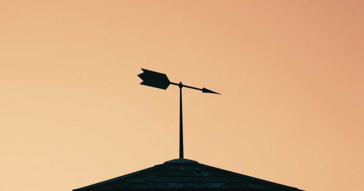 A weathervane on a rooftop against a flat white sky, showing motion without direction